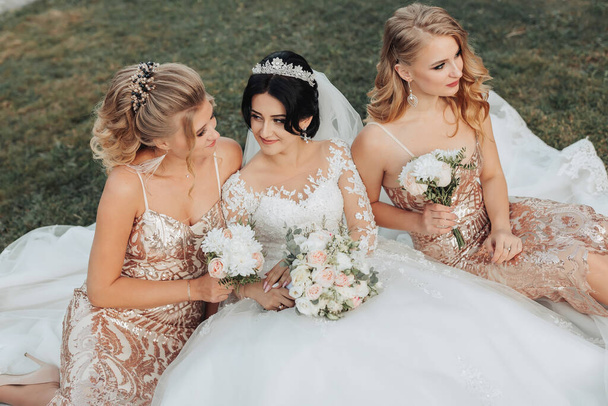 A brunette bride in a white elegant dress with a crown and her blonde friends in gold dresses pose with bouquets while sitting on the grass. Wedding portrait in nature, wedding photo in a light tone. Three beautiful women - Zdjęcie, obraz