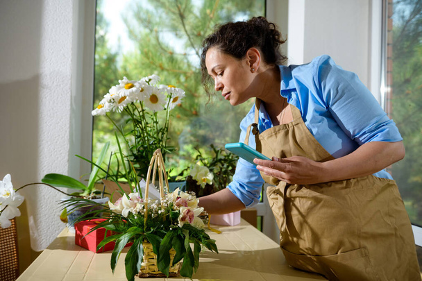 Güzel, çok ırklı kadın çiçekçi, bej önlüklü girişimci, elinde cep telefonuyla çiçeklerle bir çiçek tasarım stüdyosunda özel etkinlikler düzenliyor. - Fotoğraf, Görsel