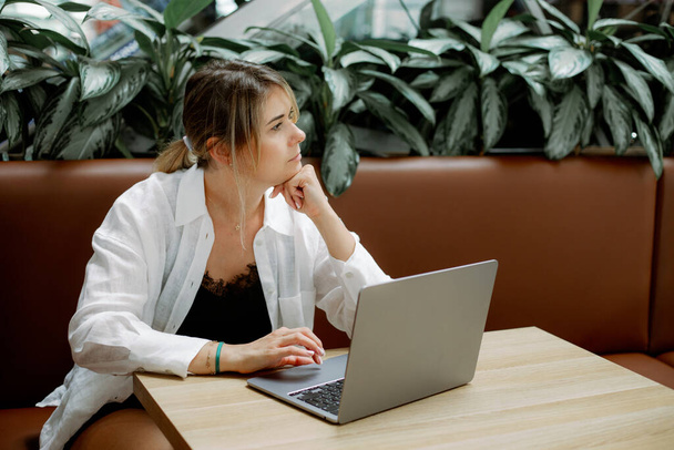Thoughtful woman with ponytail sitting on brown leather sofa at cafe table surrounded by plants, working with laptop. Freelancer in white shirt lost in thought, leaning head on hand and look aside.  - Photo, Image
