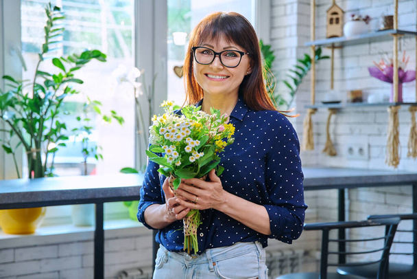 Portrait of a middle aged smiling woman with a bouquet of flowers. Happy female indoors, window table bar stools background. - Foto, afbeelding