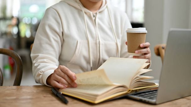 Περικοπή εικόνα μιας γυναίκας σε casual ρούχα έρευνα πληροφορίες σε ένα βιβλίο και πίνοντας καφέ, ενώ απομακρυσμένη εργασία σε μια καφετέρια το Σαββατοκύριακο. - Φωτογραφία, εικόνα