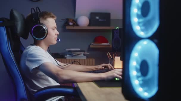 ヘッドフォンの少年は自宅でコンピュータで再生されます。ゲーマーは、グリームをオンにし、カメラに見える。PCモニターに表示されるカーレースビデオゲーム。余暇を過ごす少年。ドリーは撃たれた。サイドビュー. - 映像、動画
