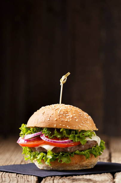 Appetizing burger with juicy beef cutlet, cheese, tomato, lettuce, onion on a wooden table and dark background. Fast food, homemade hamburger. - Foto, Imagem