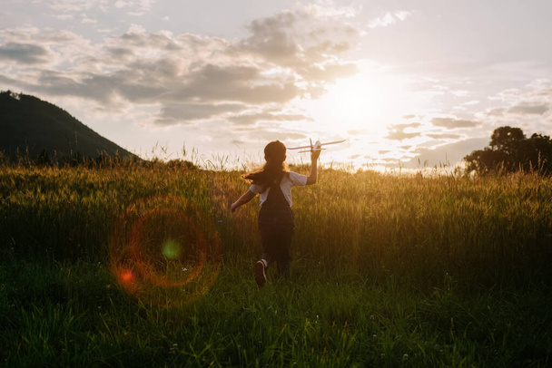 Silhouette of playful happy little girl kid run with airplane on background amazing summer sunset and majestic mountains. Dream freedom concept. Child runs on field holding in hands toy aircraft - Photo, image