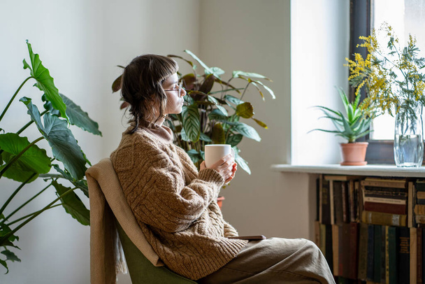 Düşünceli, yaratıcı kız elinde fincan kahveyle koltuğa oturur pencereden dışarı bakar sıcak bir evde, saksıda yeşil bitkiler olan günün tadını çıkarır. Genç kadın hayatı düşündü. Ruh sağlığı konsepti. - Fotoğraf, Görsel