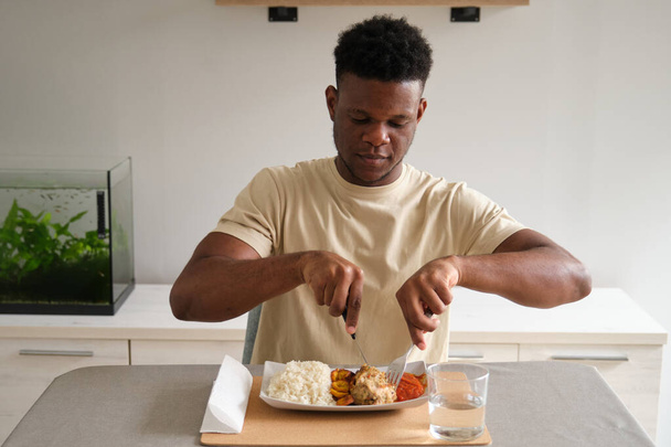Jovem negro comendo frango frito, arroz e banana com prato de molho de tomate na sala de estar. - Foto, Imagem