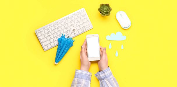 Γυναικεία χέρια που κρατούν κινητό τηλέφωνο, μικρή ομπρέλα, χάρτινο σύννεφο και πληκτρολόγιο υπολογιστή σε κίτρινο φόντο. Έννοια πρόγνωσης - Φωτογραφία, εικόνα