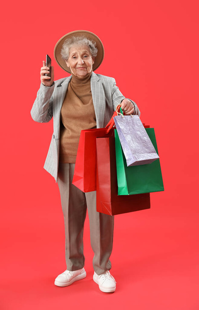Mujer mayor con teléfono móvil y bolsas de compras sobre fondo rojo - Foto, Imagen