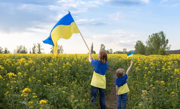 vrouw en een kind zijn gekleed in gele en blauwe T-shirts met de vlaggen van Oekraïne in hun handen tussen het koolzaadveld. onderwijs in patriottisme. Onafhankelijkheidsdag in Oekraïne. Trots om Oekraïens te zijn - Foto, afbeelding