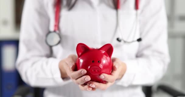 Doctor holding red piggy bank in clinic closeup 4k movie slow motion. Investment in medical education concept - Video