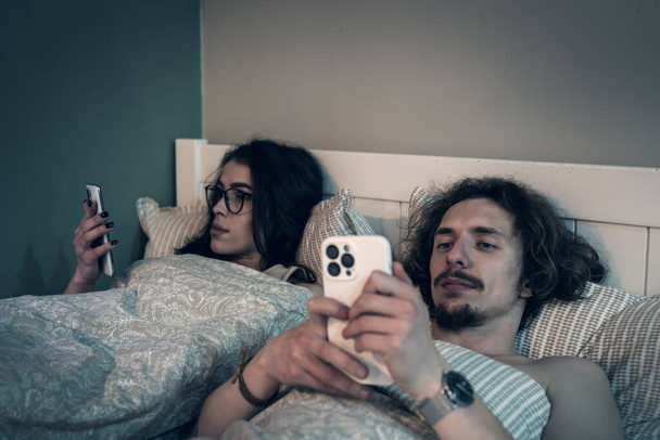 夫婦はベッドで寝そべっているが、各パートナーはスマートフォンを使う。男と女は携帯電話を見ている。ソーシャルメディア中毒、ニュースの閲覧、関係の問題. - 写真・画像