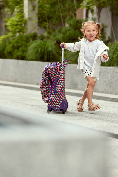 Little smiling girl, child pulling her suitcase on street, going on summer vacation with parents. Joy and happiness. Concept of family, childhood and parenthood, travelling, vacation, ad - Photo, Image