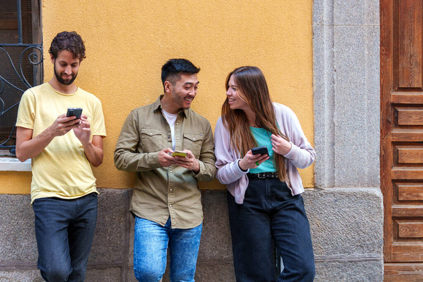 Захвачено яркое городское товарищество: трое разнообразных друзей, одетые в яркие городские наряды, погруженные в мобильный контент, прислонившись к городской стене. - Фото, изображение