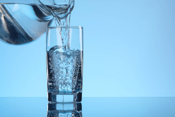 Giet water uit kan in glas op lichtblauwe achtergrond. Ruimte voor tekst - Foto, afbeelding