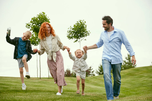Young couple, man and woman going for a walk in park with their little children, boy and girl, playing on warm summer day outdoors. Concept of family, childhood and parenthood, fun, weekends, love, ad - Photo, image