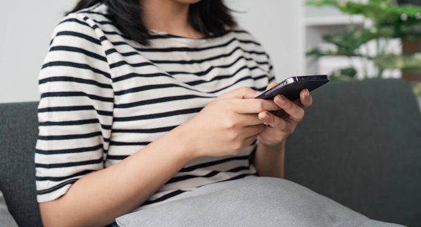 Close up of beautiful Asian woman sit on sofa hand holding mobile phone chatting with friends and playing social media. Young female looking at smartphone cellphone browsing internet at living room. - Foto, immagini