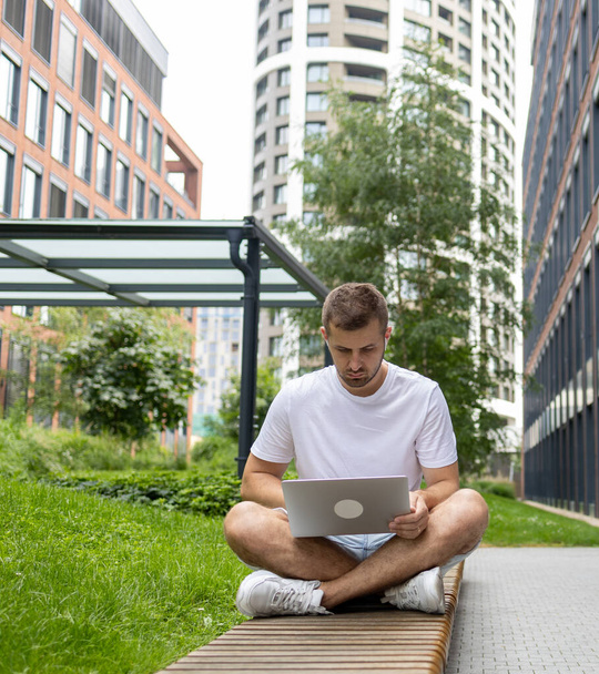 Sentado na Internet. Um estudante sério está sentado em um banco e olhando para um laptop. Fala através de fones de ouvido - Foto, Imagem