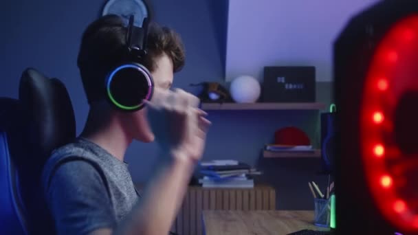 男の子はヘッドフォンに置き、自宅でコンピュータ上でゲームで再生を開始します。ゲーマーはマイクを使用してチームメイトと話します。オンラインビデオゲームライブストリーミングやサイバースポーツ選手権。サイドビュー。ドリーショット. - 映像、動画