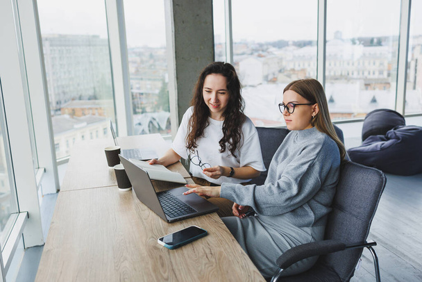 Schöne zwei junge selbstbewusste Geschäftsfrauen sitzen in einem Büroraum und arbeiten an einem Laptop. Die Arbeit der Kollegen in einem modernen Büro. - Foto, Bild