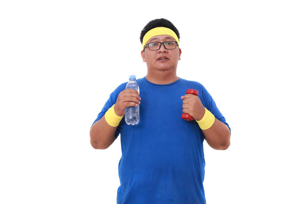 Asian overweight man in sportswear standing while holding a bottle of drink and dumbbell. Isolated - Photo, Image