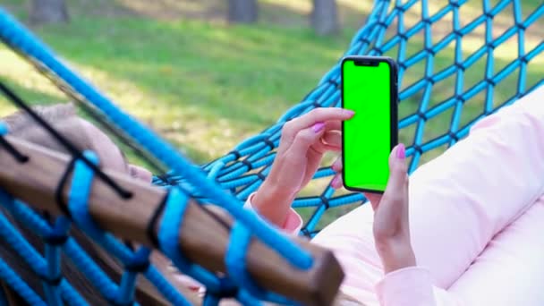 A young woman is resting in the forest, she is lying in a hammock and using a smartphone with a green screen. Outdoor recreation. Chromakey background - Materiał filmowy, wideo
