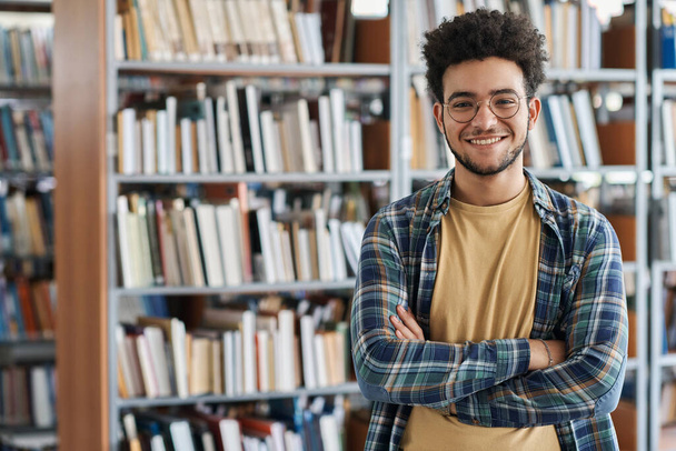Portrét dospělého studenta stojícího se založenýma rukama a usmívajícího se na kameru v knihovně na vysoké - Fotografie, Obrázek