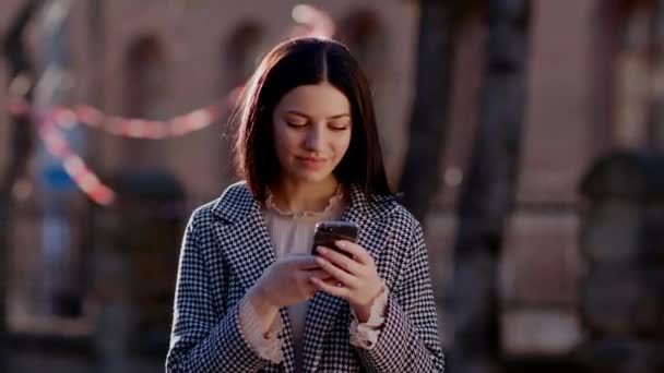 Egy csinos lány áll az utcán és gépel a telefonon. A lány az épületek és fák hátterében áll. Kiváló minőségű 4k felvételek - Felvétel, videó