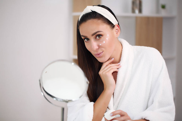 brunette in a bathrobe applied white cream on the face, skin care. - 写真・画像
