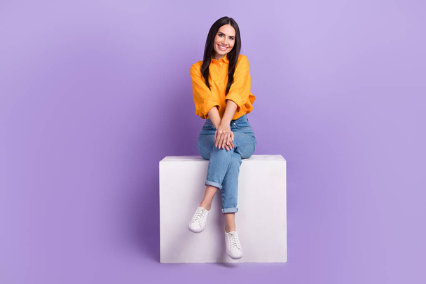 Full length photo of gorgeous friendly cute girl dressed yellow shirt denim pants sit on white cube isolated on purple color background. - Photo, image