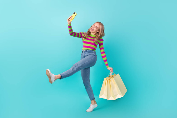Full length photo of carefree dreamy woman dressed sweater walking tacking selfie holding shoppers isolated turquoise color background. - Фото, зображення