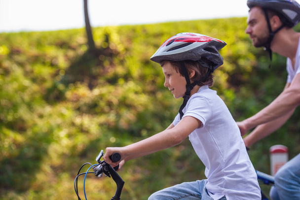 Vista lateral do menino bonito e seu pai em roupas casuais e capacetes sorrindo ao andar de bicicleta no parque. Pai em segundo plano
 - Foto, Imagem