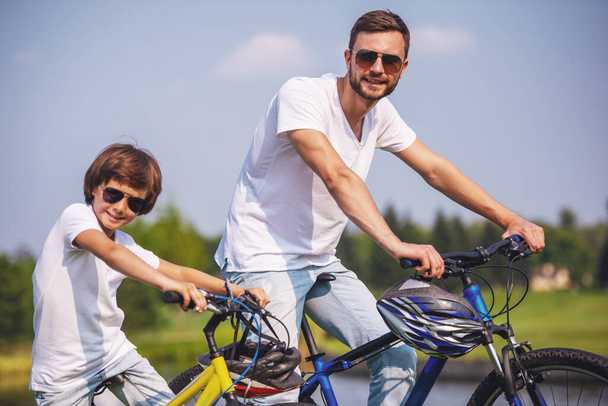 Handsome young dad and his cute little son in casual clothes and sun glasses are looking at camera and smiling while riding bikes in park - Фото, изображение