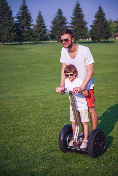Netter kleiner Junge und sein gutaussehender Vater in lässiger Kleidung und Sonnenbrille lächeln beim Segway-Fahren im Park - Foto, Bild