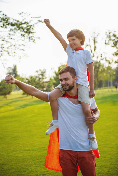 Guapo papá joven y su lindo hijo pequeño vestido como superhéroes sonríen y mantienen la mano en el puño mientras juegan en el parque
 - Foto, Imagen
