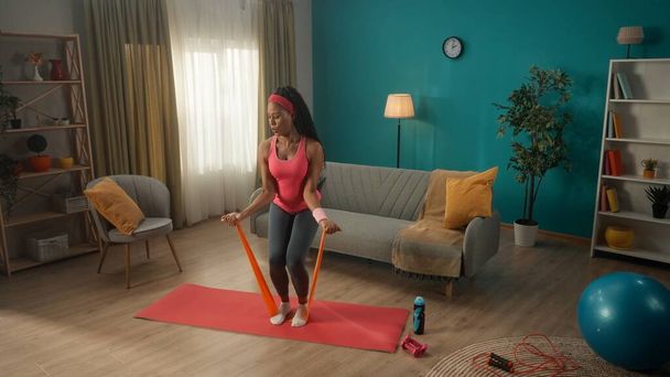 African American woman exercising with a sports elastic band at home. A young athletic woman trains her arms while standing on an elastic band. Strengthening the muscles of the biceps, triceps - Fotoğraf, Görsel