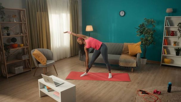 Athletic African American woman doing side bends while standing in the living room on a sports mat. A young woman watches a video lesson on a laptop and repeats the exercises. Home fitness concept - Fotó, kép