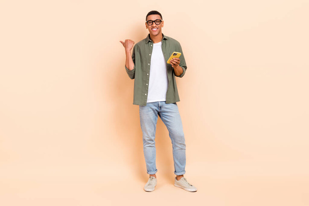 Full length photo of funky cheerful man wear khaki shirt typing modern device thumb empty space isolated beige color background. - Foto, imagen
