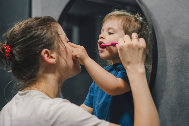 Schot van een moeder en haar dochtertje die hun tanden poetsen in de badkamer. Familie ochtend routine, tandpasta en moeder onderwijs zelfverzorging hygiëne - Foto, afbeelding