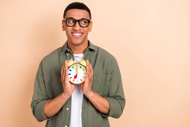 Portrait of intelligent handsome positive man wear glasses stylish shirt hold alarm look empty space isolated on beige color background. - 写真・画像