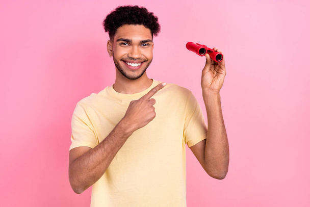 Portrait of cheerful handsome funny man with afro hairstyle earrings stubble indicating at binoculars isolated on pink color background. - Valokuva, kuva