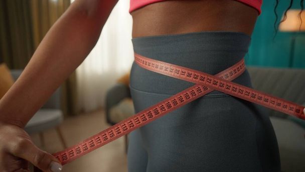 A slender African American woman measures her waist with a measuring tape. Side view of a womans thin waist wrapped in a measuring tape, close up. The concept of an ideal body, a healthy lifestyle - Foto, Imagem