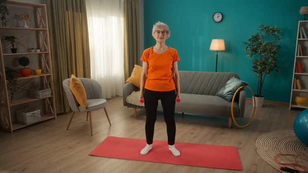 The pensioner takes care of her health, goes in for sports at home. An elderly woman trains with dumbbells, strengthening her arm muscles. A woman is standing on a sports mat, holding dumbbells in her - Foto, immagini