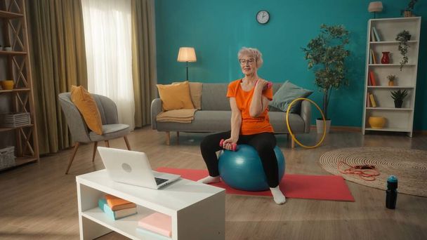 Aged woman doing sports sitting on a fitness ball in front of a laptop. An elderly woman performs a biceps exercise with dumbbells. Home fitness concept, online sports class - Foto, imagen