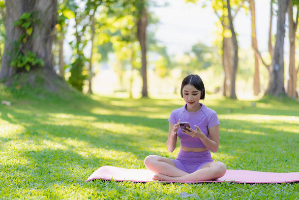 Asian woman looks at smartphone doing yoga on mat in outdoor park healthy young woman exercising yoga Yoga in natural environment for body, mind, wellness. Relax concept. - Фото, изображение