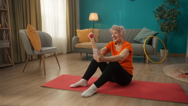 An elderly woman sits on a sports mat in the living room. A woman holds a dumbbell in one hand, exercising her hand, and a smartphone in the other hand. A woman watches a video, speaks on a video call - Φωτογραφία, εικόνα