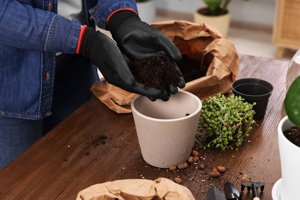 Woman in gloves filling flowerpot with soil at wooden table indoors, closeup. Transplanting houseplants - Φωτογραφία, εικόνα