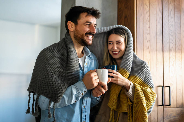 Happy young couple with hot drinks cuddling under warm blanket at home. People happiness concept. - Foto, imagen