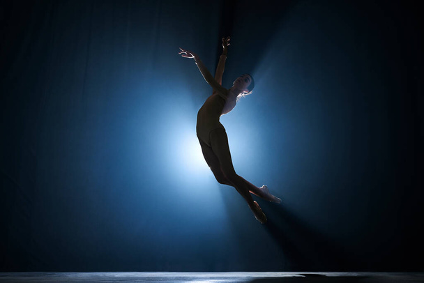Slanke, mooie, sierlijke ballerina die met schijnwerpers creatieve prestaties levert op het podium tegen donkerblauwe achtergrond. Concept van kunst, klassiek ballet, creativiteit, choreografie, schoonheid, reclame - Foto, afbeelding