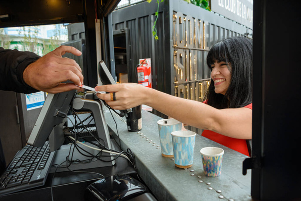 young happy venezuelan latin woman smiling handing her debit card to the waiter in the fast food cart to pay for her order, food and drink concept, copy space. - Photo, Image