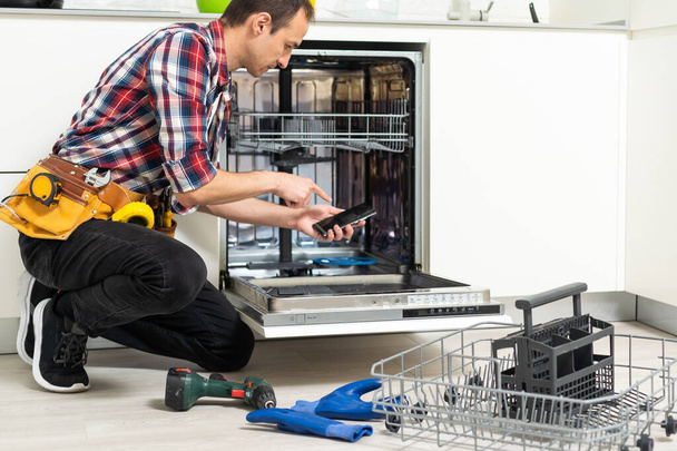 Ремонтник чинит посудомоечную машину с отверткой на кухне. Высокое качество фото - Фото, изображение